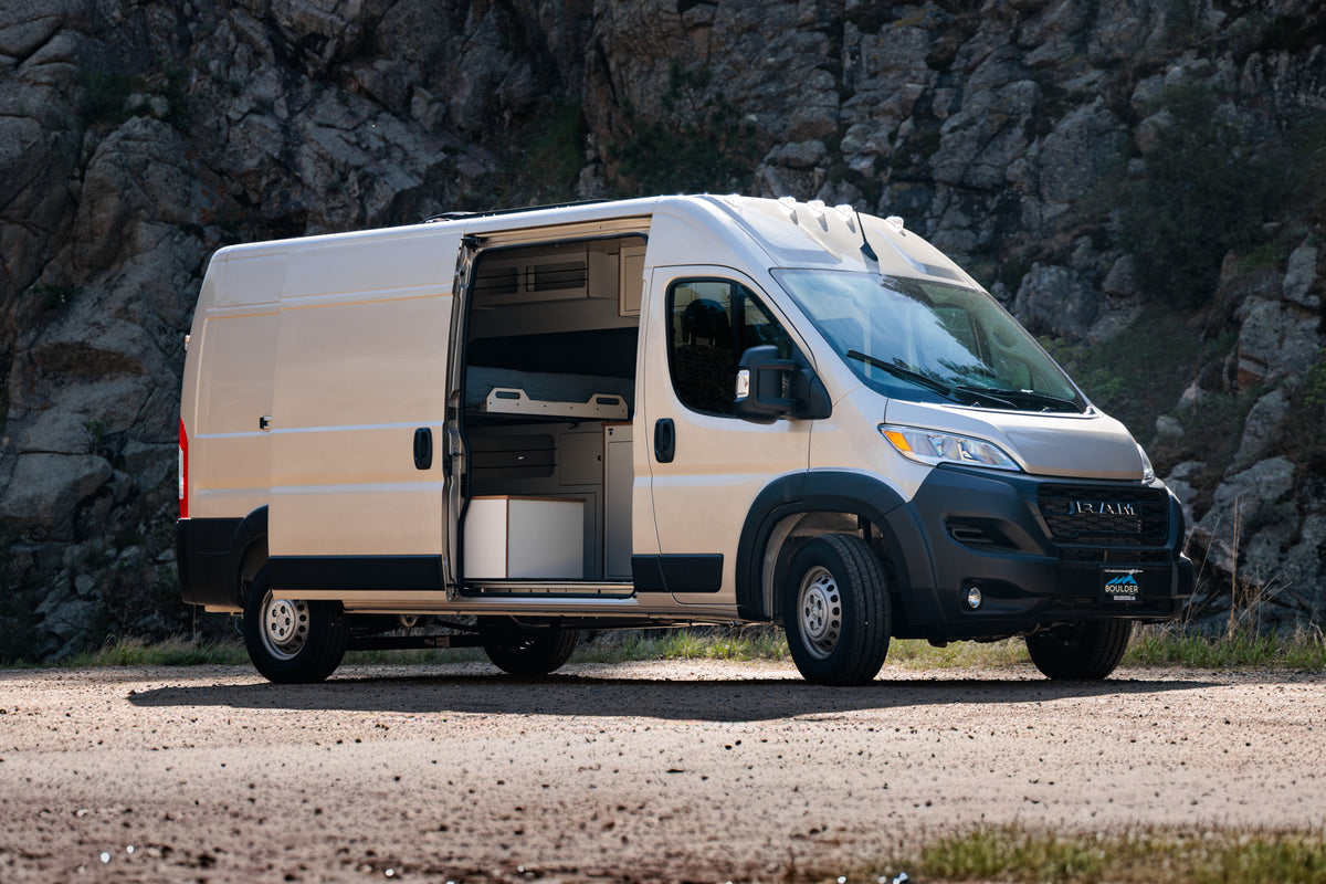 2025 RAM ProMaster Timber Camp van — sandstone exterior parked on a gravel pull-off with the sliding door open